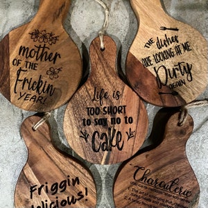 Puede incluir: Cinco tablas de servir de madera con diferentes refranes humorísticos grabados en ellas. Los refranes incluyen "¡Madre del año del frickin!" "La vida es demasiado corta para decir que no al pastel" "Los platos me están mirando sucio de nuevo" "¡Friggin delicioso!" y "Charcutería".