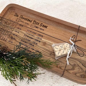 Peut inclure: Plateau de jeu de dés Ten Thousand en bois avec règles et dés gravés. Le plateau rectangulaire a des coins arrondis et un grain de bois foncé. Un petit sac de dés est attaché au plateau avec de la ficelle. De la verdure décorative est dans le coin.