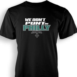 以下が含まれることがあります： 白とティール色の文字で「WE DON'T PUNT IN PHILLY」と書かれた黒いTシャツ。シャツにはテキストの下にフットボールをテーマにしたグラフィックが描かれています。スポーツファン向けのカジュアルなTシャツです。