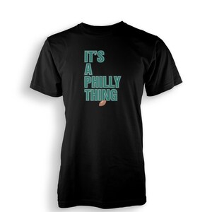 以下が含まれることがあります： 緑色の「It's a Philly Thing」の文字と茶色のフットボールのグラフィックがプリントされた黒いTシャツ