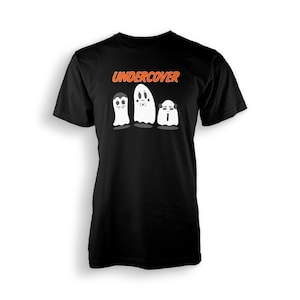 Op de afbeelding: Zwart t-shirt met het woord "UNDERCOVER" in oranje boven drie cartoongeesten. De geesten hebben verschillende uitdrukkingen en accessoires, waaronder een vlinderdas en vampierkenmerken.