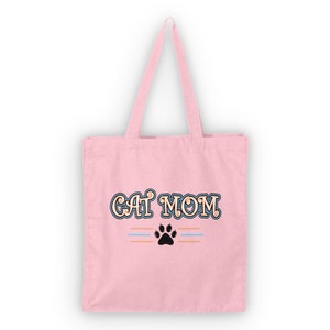 Könnte beinhalten: Eine rosa Canvas-Tragetasche mit dem Text "CAT MOM" in einer dekorativen Schriftart. Der Text ist blau umrandet und hat eine braune Füllung. Unter dem Text befindet sich ein schwarzer Pfotenabdruck mit zwei dünnen blauen und orangefarbenen Streifen über dem Pfotenabdruck.