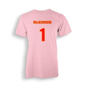 以下が含まれることがあります： ピンクのTシャツに「MCKINNON」という名前と「1」という数字が、黄色の輪郭で囲まれた赤色で印刷されています。