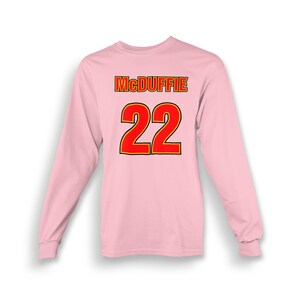 Può includere: Maglietta rosa a maniche lunghe con il nome "McDuffie" e il numero "22" stampato sul retro in rosso con un contorno giallo.