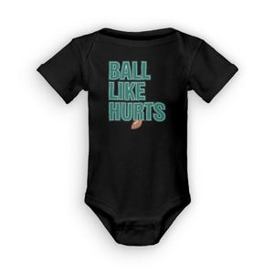 Puede incluir: Mono negro para bebé con un gráfico verde que dice "BALL LIKE HURTS" con un gráfico de fútbol americano marrón debajo del texto.