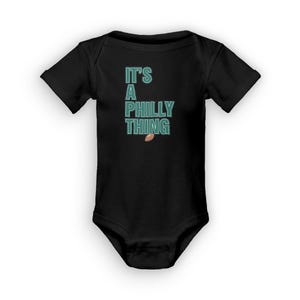 以下が含まれることがあります： 黒のベビーロンパースに「It's a Philly Thing」という文字がターコイズグリーンで書かれており、茶色のフットボールのグラフィックが描かれています。
