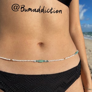 Puede incluir: Una cadena de cintura de cuentas blancas con detalles turquesa y dorados. La cadena se lleva en la cintura de una persona. El fondo es una playa.