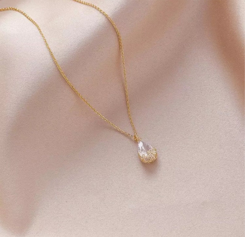 May include: A delicate gold chain necklace with a teardrop-shaped crystal pendant. The crystal is clear and has a faceted design.