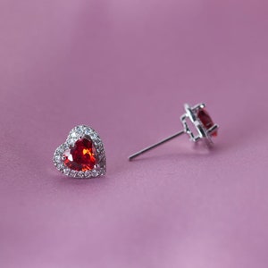 May include: A pair of silver heart-shaped earrings with a red gemstone in the centre and a halo of smaller clear gemstones.