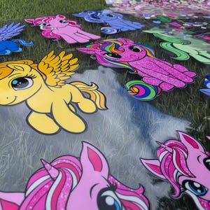 May include: A collection of colorful pony and unicorn stickers on a transparent sheet. The designs feature various characters in shades of yellow, pink, blue, and green, with some elements displaying a glittery texture and rainbow-colored manes.
