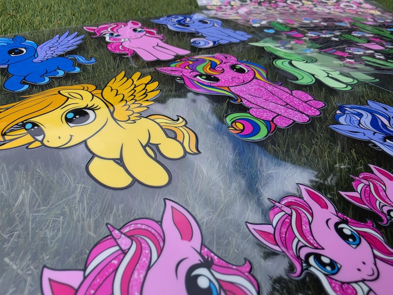 May include: A collection of colorful pony and unicorn stickers on a transparent sheet. The designs feature various characters in shades of yellow, pink, blue, and green, with some elements displaying a glittery texture and rainbow-colored manes.
