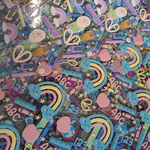 May include: A collection of school-themed stickers featuring pastel-colored rainbows with clouds, rulers, pencils, apples, scissors, and the letters ABC and 123. The designs include various educational icons in shades of blue, pink, yellow, and purple.