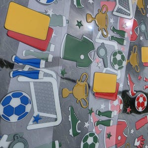 May include: A collection of soccer-themed stickers on a clear sheet, featuring blue and white soccer balls, gold trophies, green jerseys, red and yellow penalty cards, whistles, soccer goals, and green and white cleats, accented with small stars.