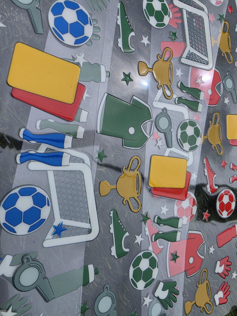 May include: A collection of soccer-themed stickers on a clear sheet, featuring blue and white soccer balls, gold trophies, green jerseys, red and yellow penalty cards, whistles, soccer goals, and green and white cleats, accented with small stars.