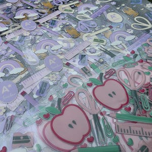 May include: A collection of school-themed stickers featuring purple rainbows, pink apples, scissors, rulers, pencils, and circular labels with the text A+ teacher. The designs include various stationery items in soft pastel shades of pink, purple, and green.