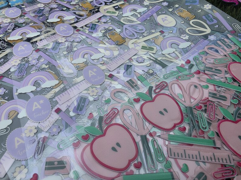 May include: A collection of school-themed stickers featuring purple rainbows, pink apples, scissors, rulers, pencils, and circular labels with the text A+ teacher. The designs include various stationery items in soft pastel shades of pink, purple, and green.