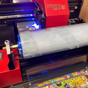 May include: A red and black industrial UV printer featuring a large roll of transparent film. The machine has a bright red housing with metallic text and a blue light illuminating the printing area, while a printed sheet with colorful cartoon characters.