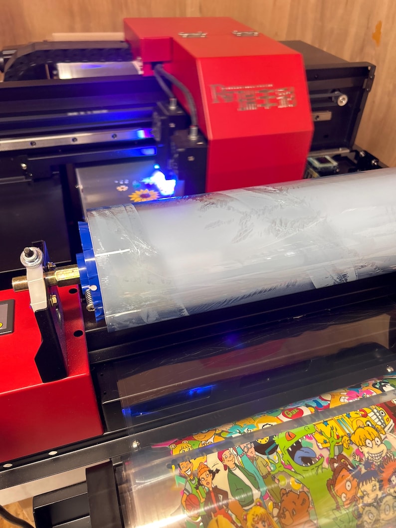 May include: A red and black industrial UV printer featuring a large roll of transparent film. The machine has a bright red housing with metallic text and a blue light illuminating the printing area, while a printed sheet with colorful cartoon characters.
