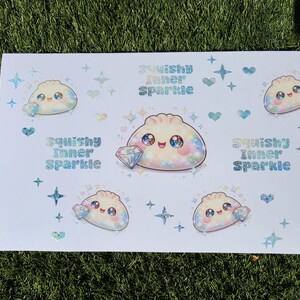 May include: A white sticker sheet featuring multiple illustrations of a smiling, round dumpling character with large, sparkling eyes. Each character holds a diamond. The text "Squishy Inner Sparkle" appears in a blue, glittery font alongside small stars and hearts.