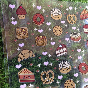 May include: A clear sticker sheet featuring various baked goods including cupcakes, donuts, waffles, pancakes, croissants, pretzels, cookies, and cake slices. Each food item has a smiling face, interspersed with small pink hearts and white stars.