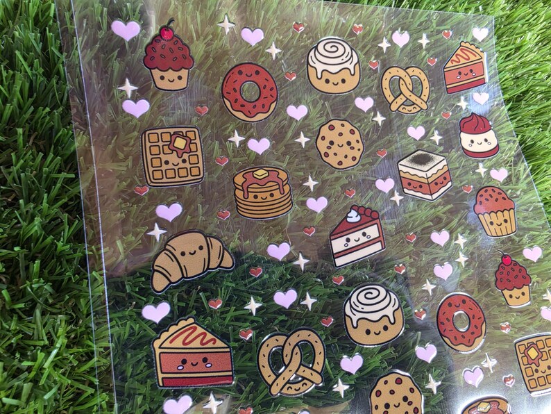 May include: A clear sticker sheet featuring various baked goods including cupcakes, donuts, waffles, pancakes, croissants, pretzels, cookies, and cake slices. Each food item has a smiling face, interspersed with small pink hearts and white stars.
