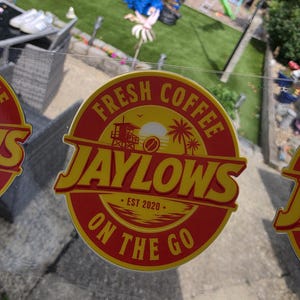 May include: A circular sticker with a red background and yellow border featuring the text FRESH COFFEE JAYLOWS ON THE GO and EST 2020. The graphic includes a lifeguard tower, a sun, palm trees, and a coffee bean icon in the center.