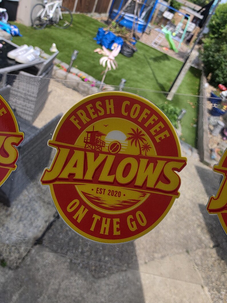 May include: A circular sticker with a red background and yellow border featuring the text FRESH COFFEE JAYLOWS ON THE GO and EST 2020. The graphic includes a lifeguard tower, a sun, palm trees, and a coffee bean icon in the center.