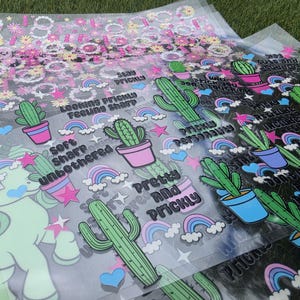 May include: A collection of clear transfer sheets featuring printed designs of green cacti in pink and blue pots, colorful rainbows, pink stars, and blue hearts. Text includes phrases like Stay Prickly, Looking Prickly Feeling Sharp, and Pretty and Prickly.