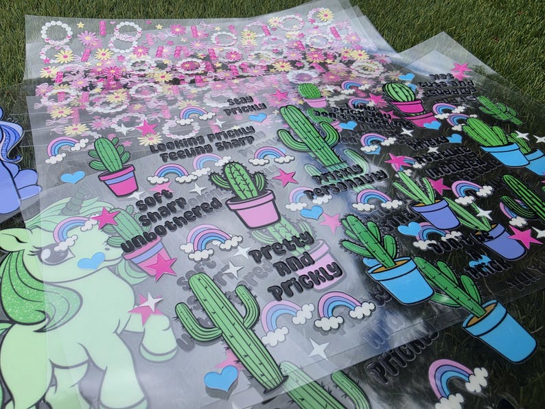May include: A collection of clear transfer sheets featuring printed designs of green cacti in pink and blue pots, colorful rainbows, pink stars, and blue hearts. Text includes phrases like Stay Prickly, Looking Prickly Feeling Sharp, and Pretty and Prickly.