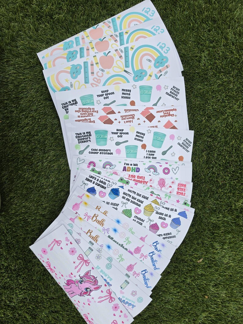 May include: A collection of rectangular sticker sheets featuring various colorful designs, including rainbows, apples, ice cream cups, cupcakes, and a pink unicorn. The sheets contain printed text phrases like 'Brain Freeze builds character' and 'I'm a bit ADHD'.