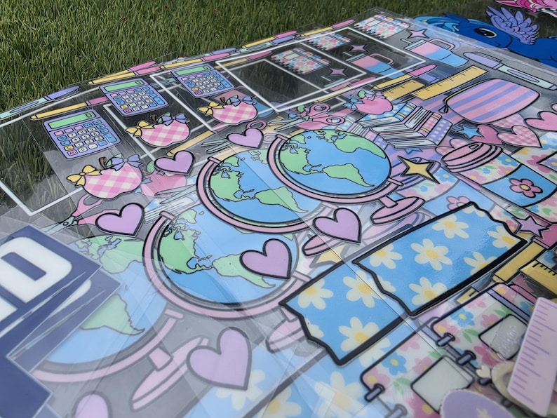 May include: A collection of school-themed decorative cutouts featuring illustrations of globes, calculators, apples, rulers, and hearts in a pastel color palette of light blue, pink, and yellow, printed on clear plastic sheets with a glossy finish.