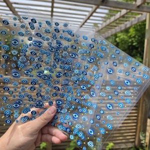 May include: A transparent sheet featuring a repeating pattern of blue and white evil eye symbols interspersed with small gold star icons. The design consists of multiple eye motifs in varying sizes, creating a dense, decorative graphic print on clear film.