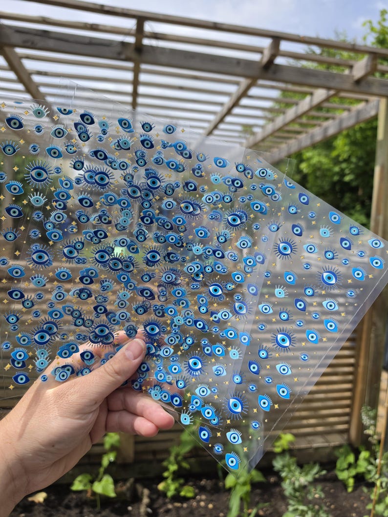 May include: A transparent sheet featuring a repeating pattern of blue and white evil eye symbols interspersed with small gold star icons. The design consists of multiple eye motifs in varying sizes, creating a dense, decorative graphic print on clear film.