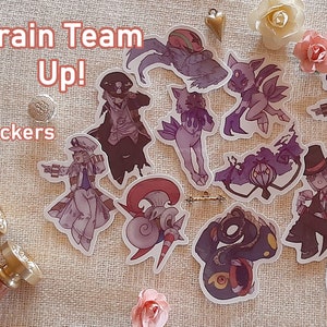 May include: A set of 12 stickers featuring cartoon characters with a purple and white color scheme. The stickers are on a beige fabric background. The text "Train Team Up! Stickers" is visible in the top left corner of the image.