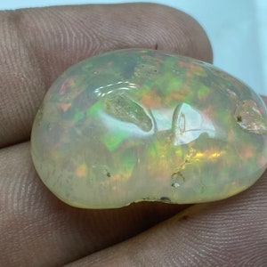 May include: A white opal gemstone with green, orange, and pink iridescent flashes. The opal is smooth and polished, with a rounded shape.
