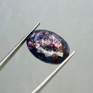 May include: A dark blue oval-shaped gemstone with iridescent inclusions of pink, gold, and purple. The gemstone is held by a pair of tweezers.
