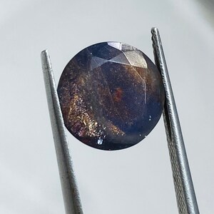 Pu&ograve; includere: Una pietra preziosa rotonda di colore blu scuro con macchie dorate e argentate. La pietra preziosa &egrave; tenuta da una pinzetta.