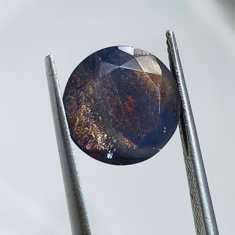 Pu&ograve; includere: Una pietra preziosa rotonda di colore blu scuro con macchie dorate e argentate. La pietra preziosa &egrave; tenuta da una pinzetta.