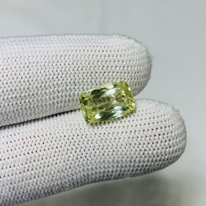 May include: A light yellow gemstone with a rectangular shape. The gemstone is cut and polished, and it has a slight shimmer.