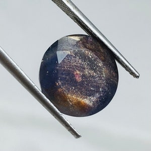 Pu&ograve; includere: Una pietra preziosa rotonda e sfaccettata di colore blu scuro, viola e oro. La pietra preziosa ha un aspetto maculato ed &egrave; tenuta da una pinzetta.