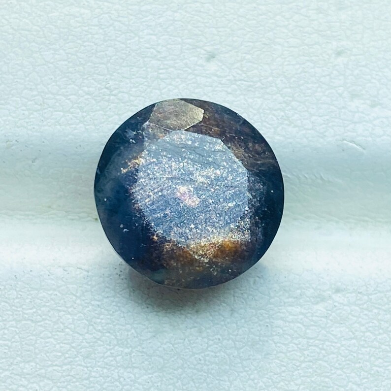 Pu&ograve; includere: Una pietra preziosa rotonda e sfaccettata di colore blu scuro e oro. La pietra preziosa ha una finitura scintillante e iridescente.