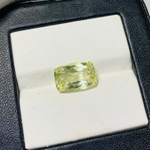 May include: A pale yellow gemstone with a rectangular cut, displayed in a white ring box.