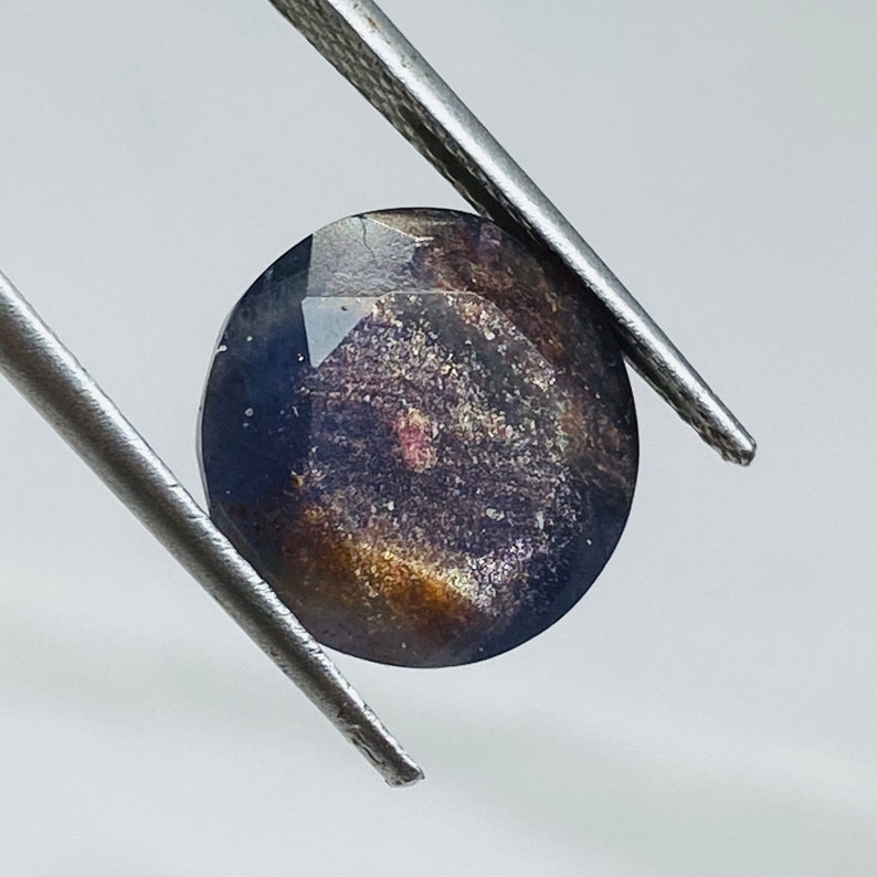 Pu&ograve; includere: Una pietra preziosa rotonda e sfaccettata di colore blu scuro, viola e oro. La pietra preziosa ha un aspetto maculato.