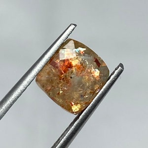 Könnte beinhalten: Ein goldener Sunstone-Edelstein mit einem Kissen-Schliff und einer rotbraunen Farbe. Der Stein wird in einer Pinzette gehalten.