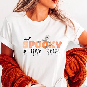 Può includere: Maglietta bianca con un design di tecnico radiologo spettrale. Il design presenta uno scheletro che tiene una radiografia, un pipistrello e le parole "Spooky X-Ray Tech" in lettere arancioni e nere che gocciolano.