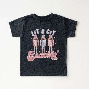 May include: A dark grey t-shirt with a festive design featuring three nutcracker figures and the text "Let's Get Crackin'".