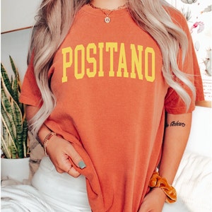 May include: A woman wearing a burnt orange t-shirt with the word "POSITANO" printed in yellow. She is wearing white trousers and has a gold chain necklace on.
