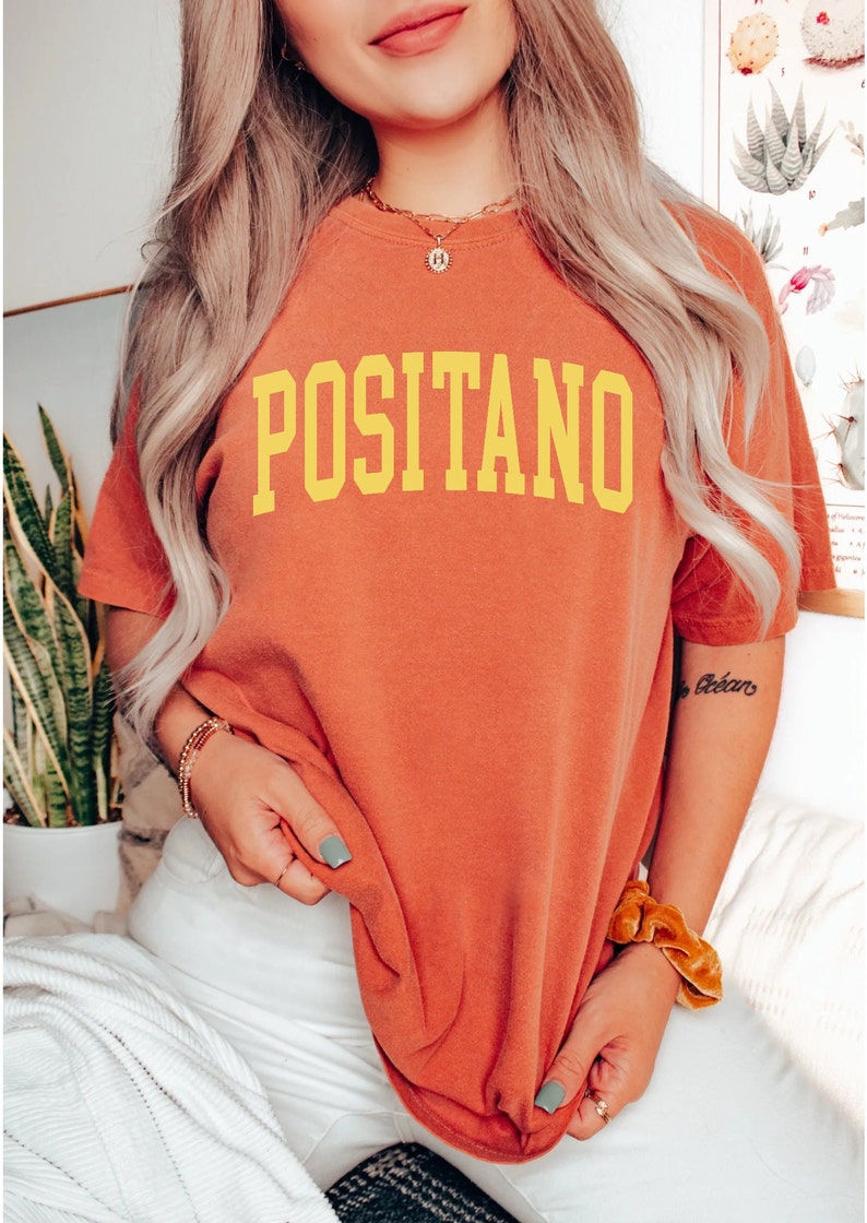 May include: A woman wearing a burnt orange t-shirt with the word "POSITANO" printed in yellow. She is wearing white trousers and has a gold chain necklace on.
