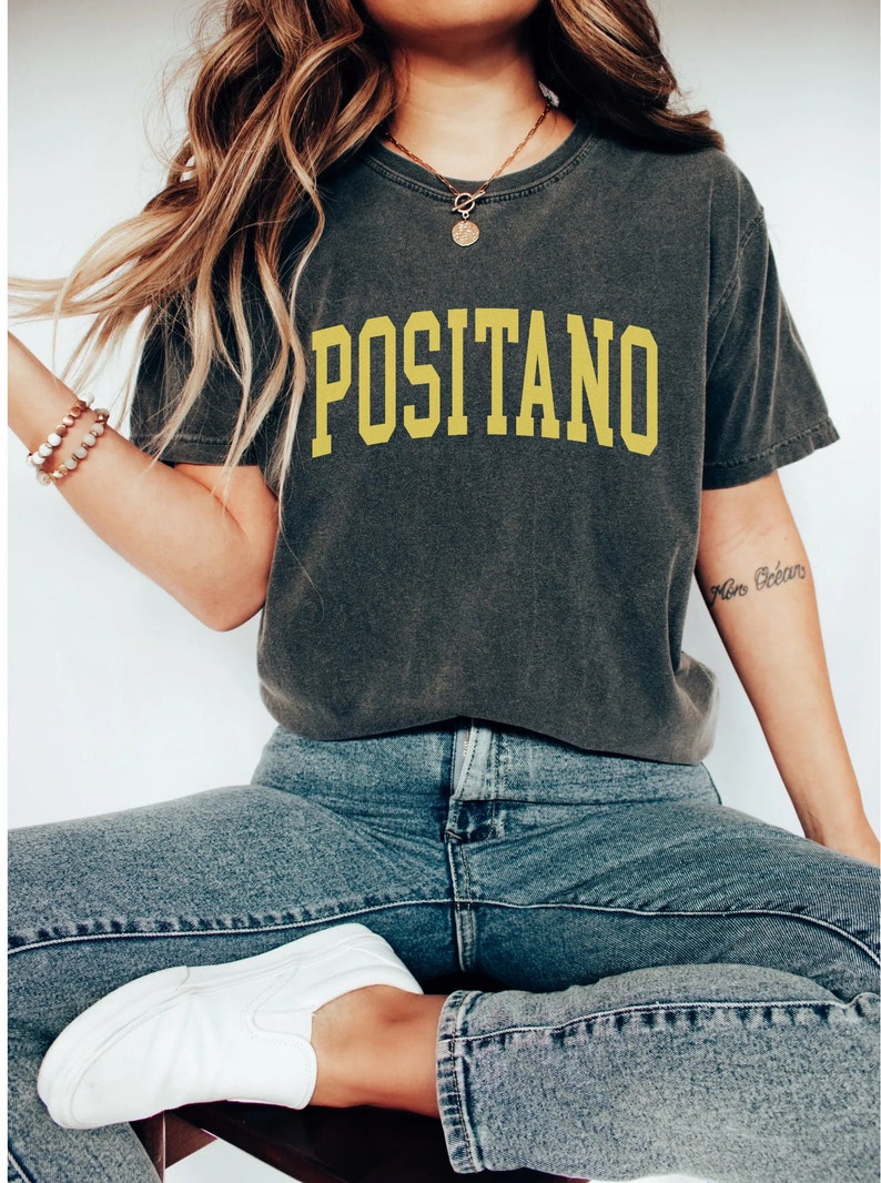 May include: A dark grey t-shirt with the word "POSITANO" printed in yellow on the front. The shirt is being worn by a person with long brown hair and blue jeans.