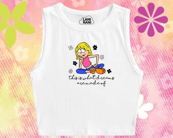May include: White vest top featuring a cartoon girl illustration, flowers, and the text "this is what dreams are made of." The top has a round neck and a small logo at the top.